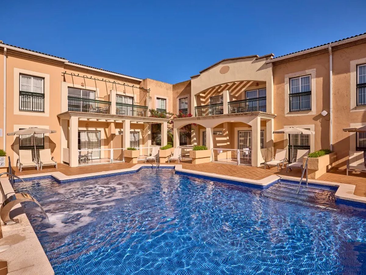 A rectangular pool between two-storey apartment blocks, with ground-floor terraces, parasols and sun loungers at the water's edge.