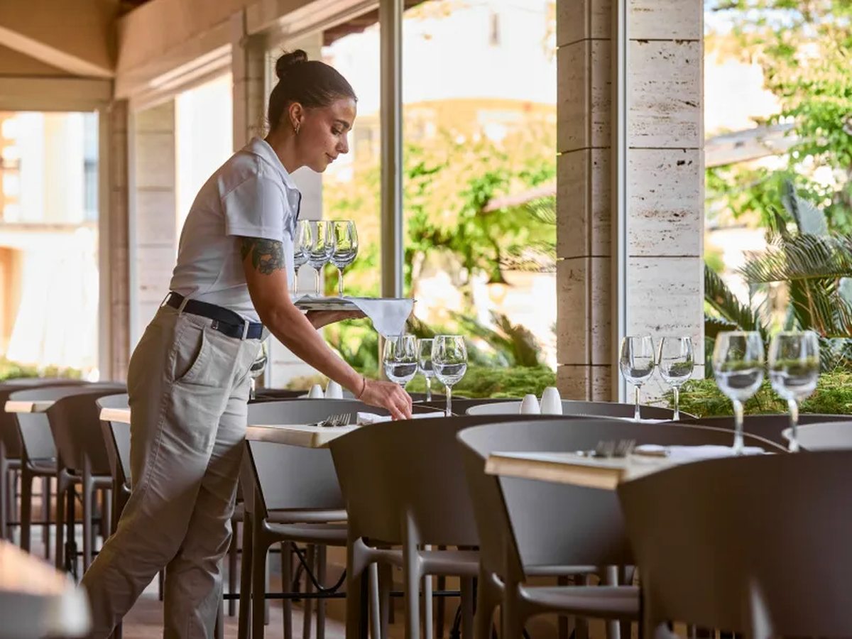 A covered outdoor dining terrace with laid tables and wine glasses, stone columns and greenery beyond, and a waitress setting a place.