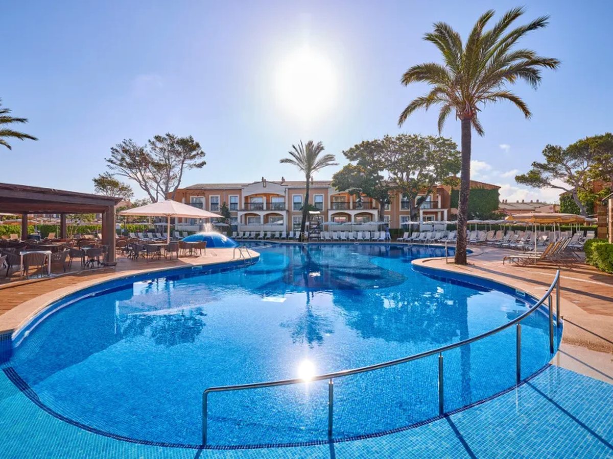 A large curved pool in bright sun with steps and a handrail, apartment blocks and palms behind and the pool bar terrace to one side.