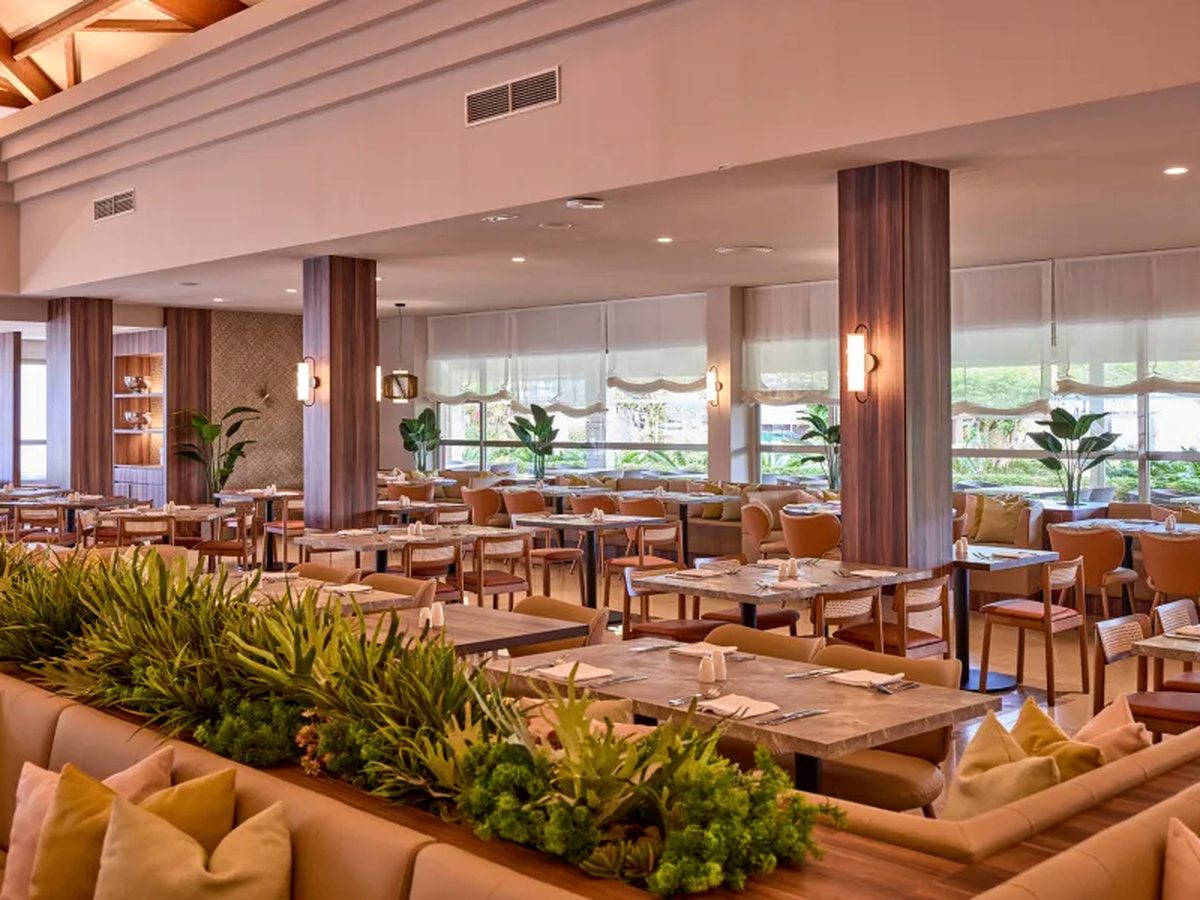 A restaurant dining room with marble-topped tables, peach upholstered chairs and banquettes, planted dividers and tall windows with pale blinds.