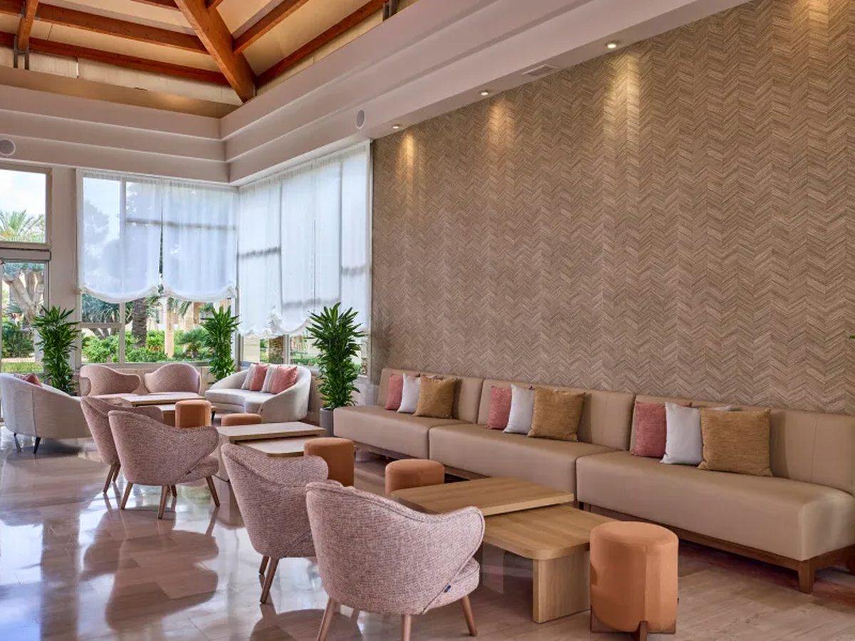 A high-ceilinged lounge with timber roof beams, a long banquette with pink and gold cushions, pink tub chairs, low wooden tables and tall windows onto the gardens.