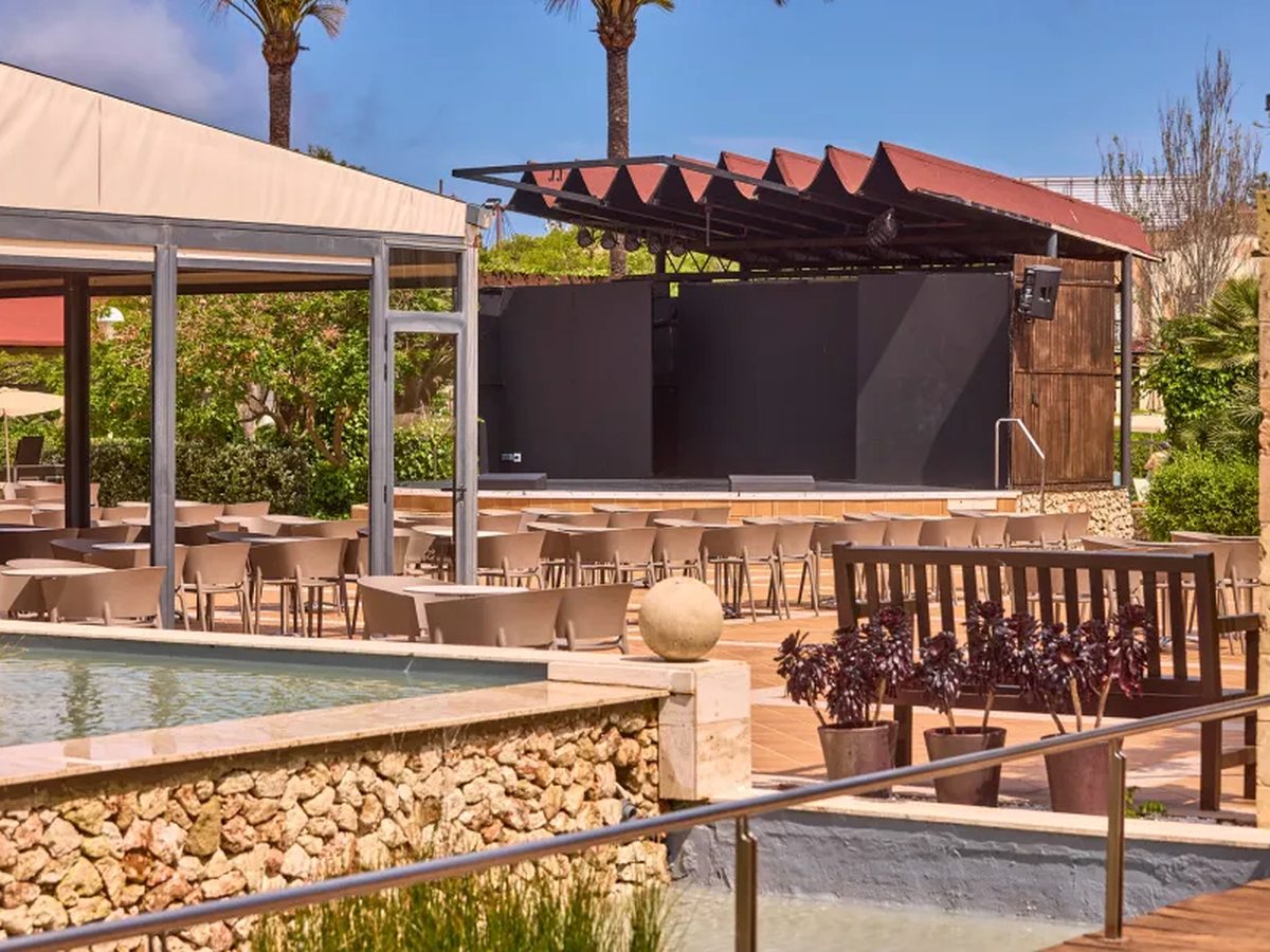 An outdoor stage with a red zigzag canopy facing rows of chairs and tables on a paved terrace, with palms and a water channel in front.