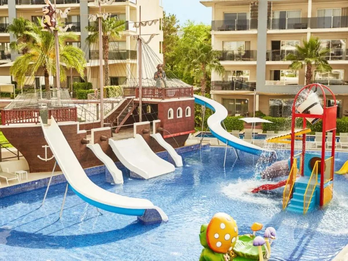 A wooden pirate ship in a shallow pool with three white slides running from it, a red and yellow splash tower to one side and suite balconies behind.