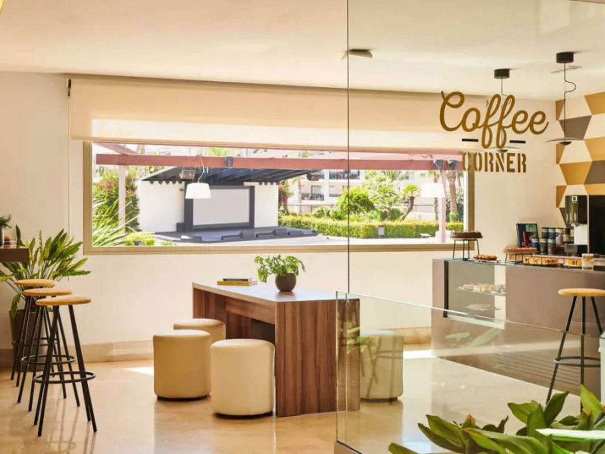 A small cafe counter with a glass pastry display and coffee machine, bar stools at a wooden table and a Coffee Corner sign on the wall.