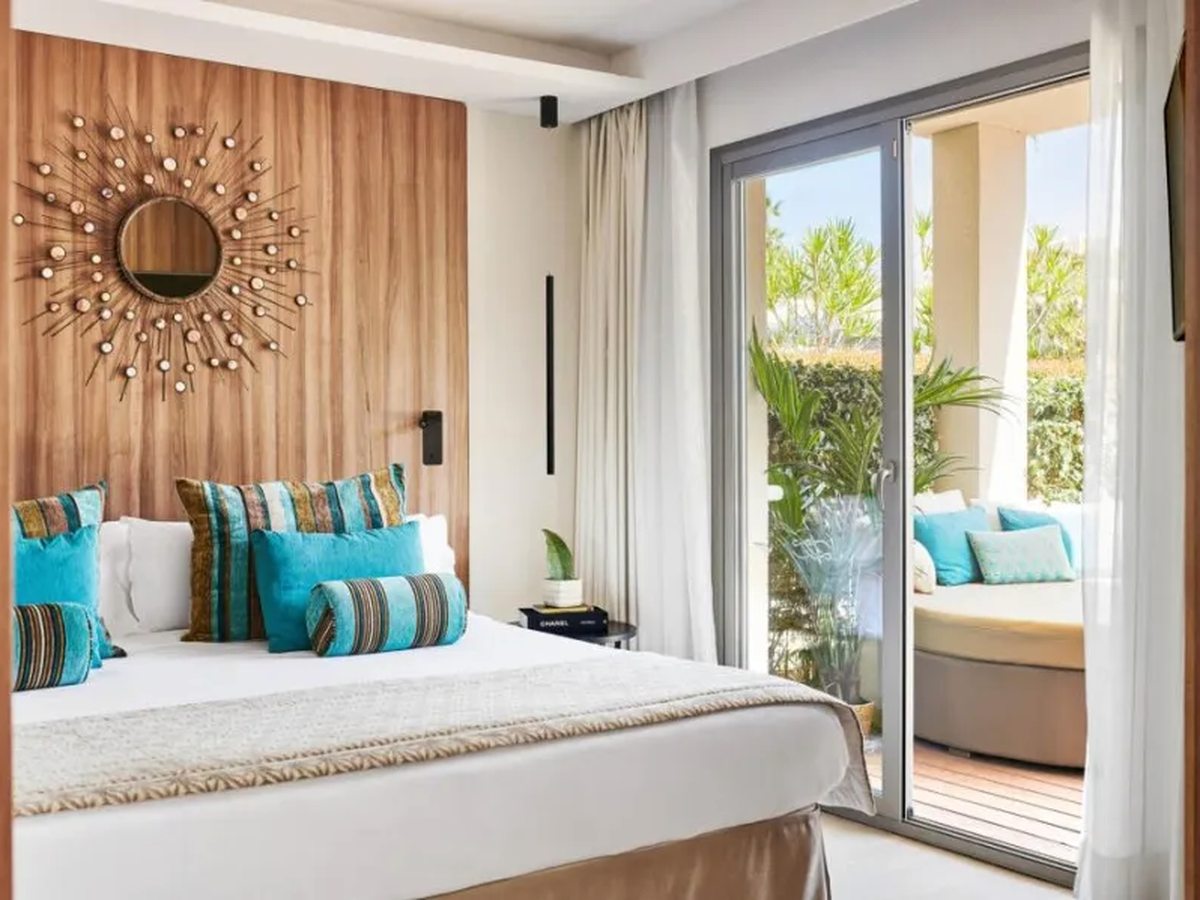 A wide bed with turquoise cushions against a wooden panelled wall with a sunburst mirror, and sliding doors open to a terrace daybed.