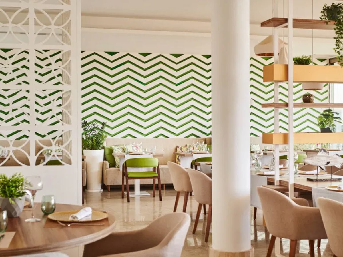 A bright dining room with a green and white zigzag tiled wall, white lattice screens, round wooden tables and cream and lime chairs.