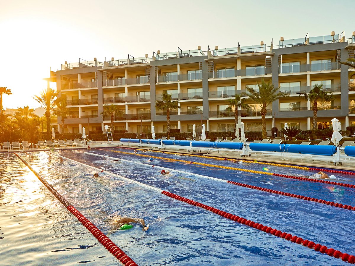Swimmers doing lengths in a pool marked out with red and yellow lane ropes, with low sun behind the accommodation block.