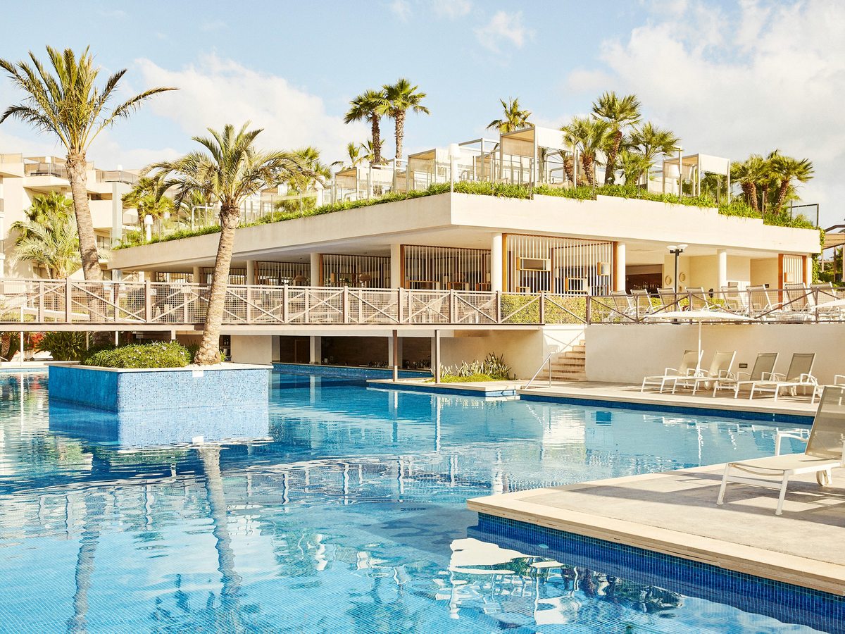 A large blue pool below a raised restaurant terrace, with sunloungers along the far edge and palms rising behind.