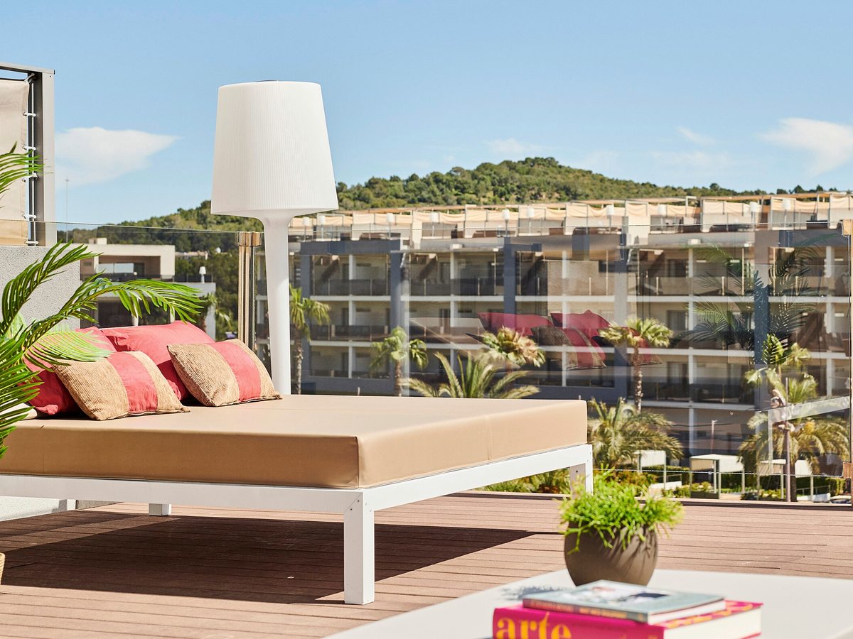 A cushioned daybed with pink and gold cushions on a timber roof deck, a tall white lamp beside it and the resort and a green hill behind.