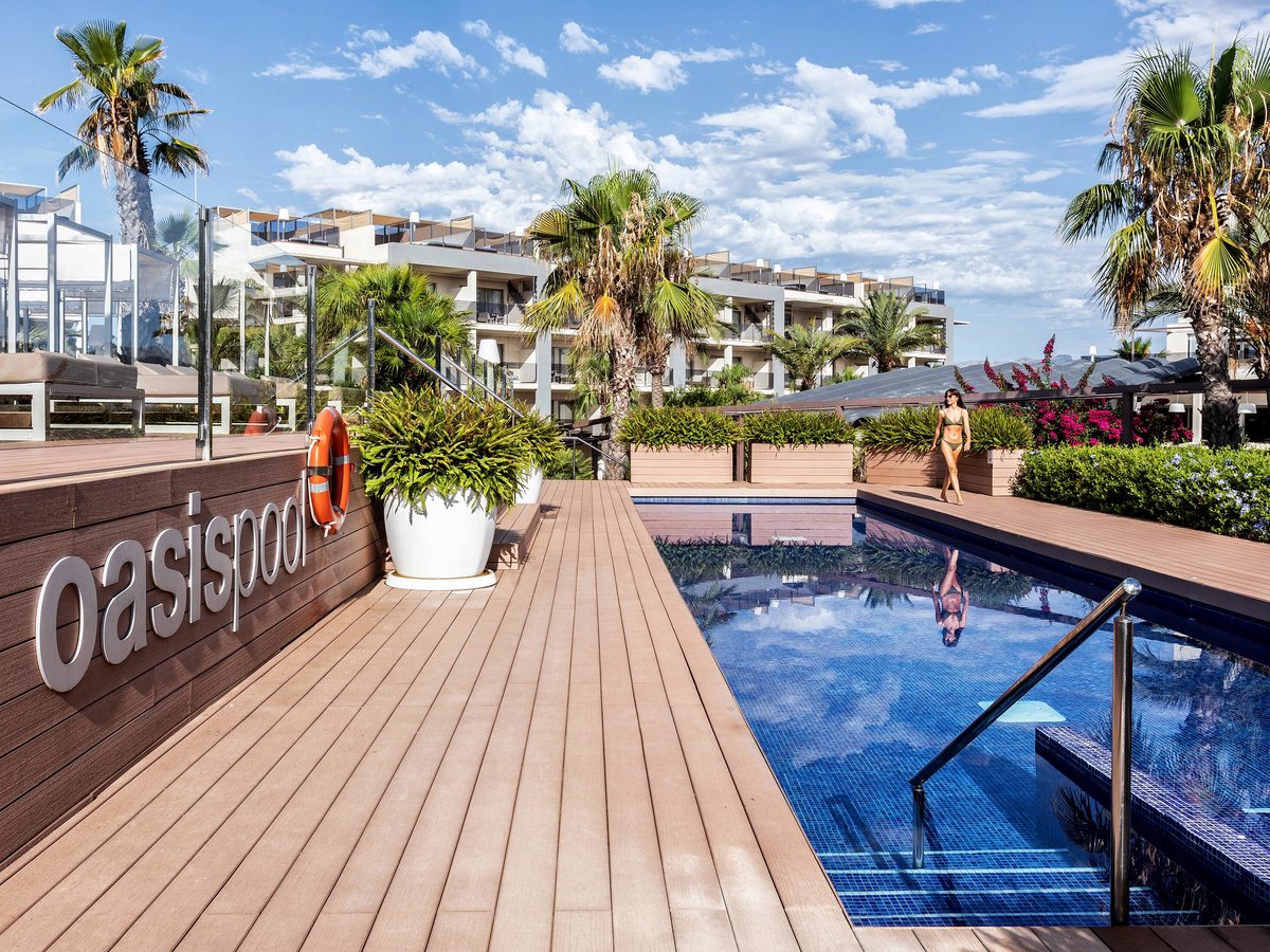 A narrow pool beside a timber deck with Oasis Pool lettering on the wall, a lifebuoy and planted tubs, and the suite blocks behind.