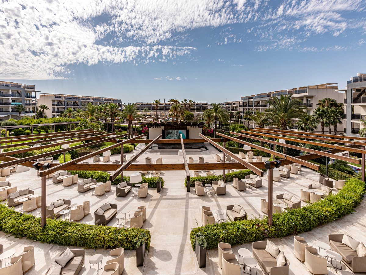 A wide open-air terrace of cream sofas and low tables in curved rows under timber pergolas, facing a small stage with a screen.