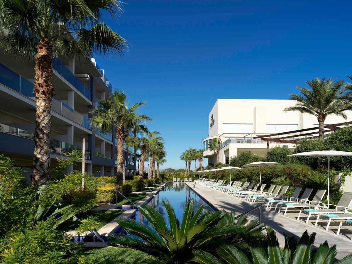 A long narrow pool between palms and hedges, with rows of sunloungers and parasols down one side and a white building at the end.