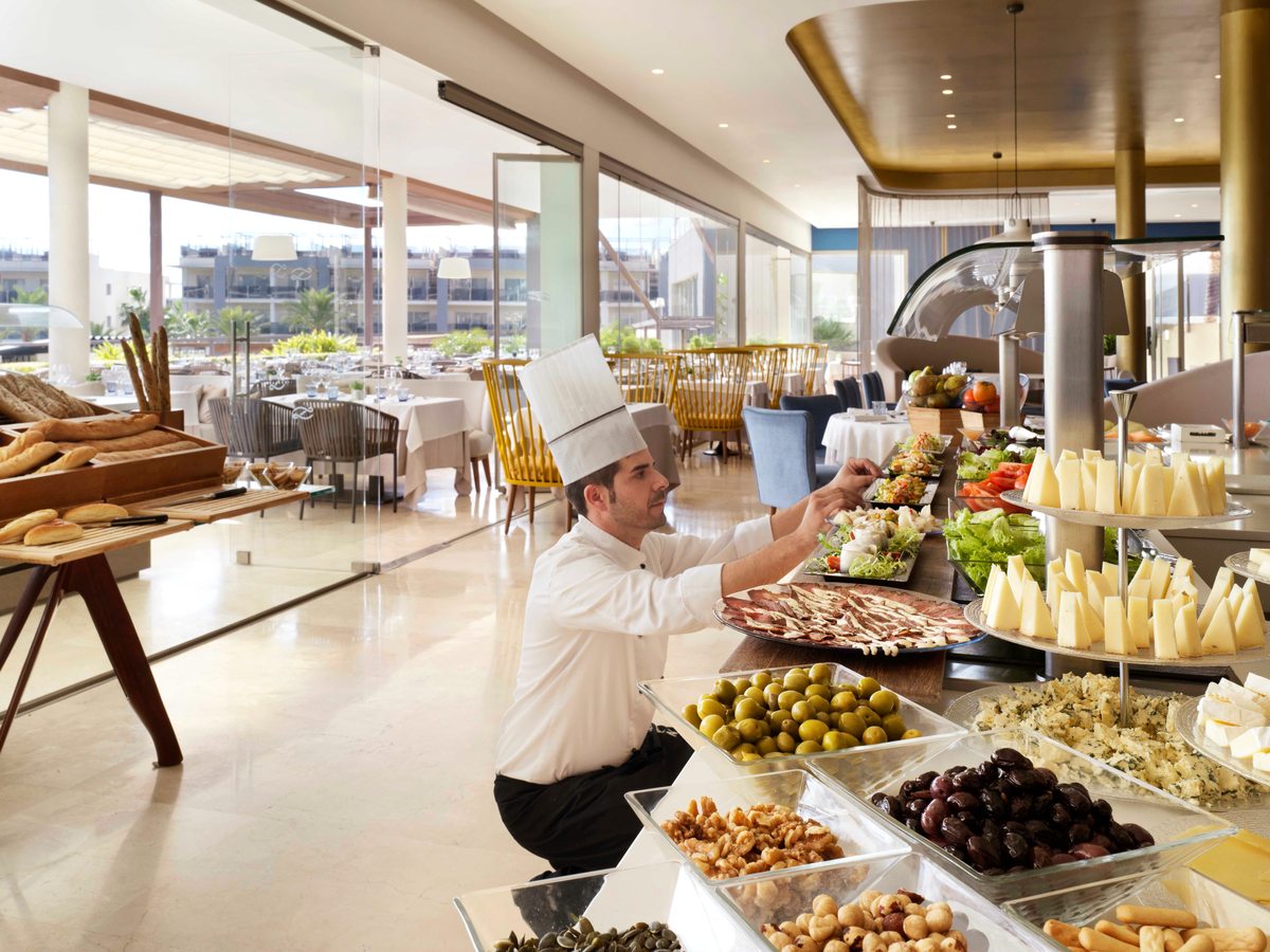 A chef in whites arranging plates of salad and cured meats at a buffet counter stacked with cheeses, olives and nuts, with laid tables behind.