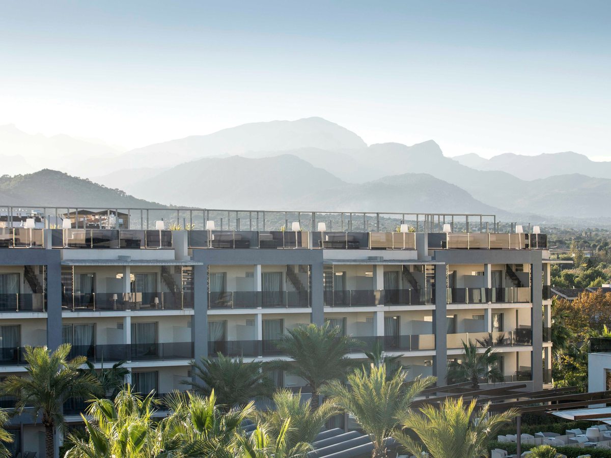 Four storeys of balconies on a white resort block, palm trees in front and hazy blue mountain ridges rising behind.