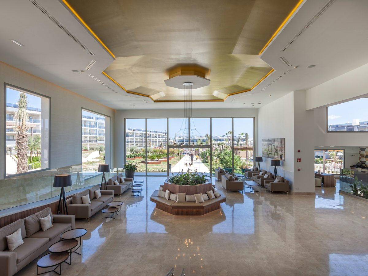 A bright marble-floored lobby with beige sofas, a circular planted bench in the middle and a glazed wall looking down the central gardens.