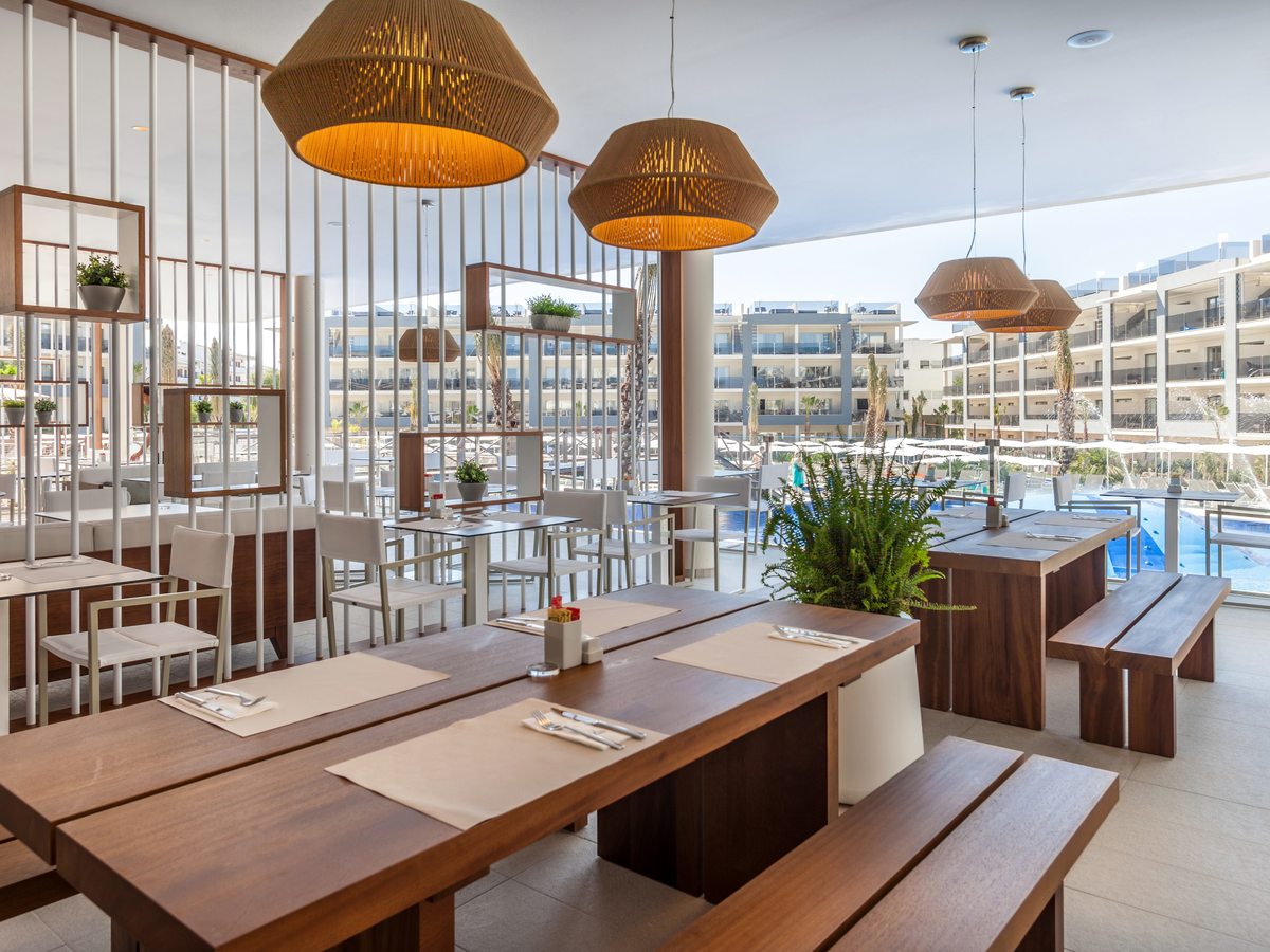 Long wooden bench tables under woven pendant lamps, with a wall of glass looking out to the pool and the suite blocks.