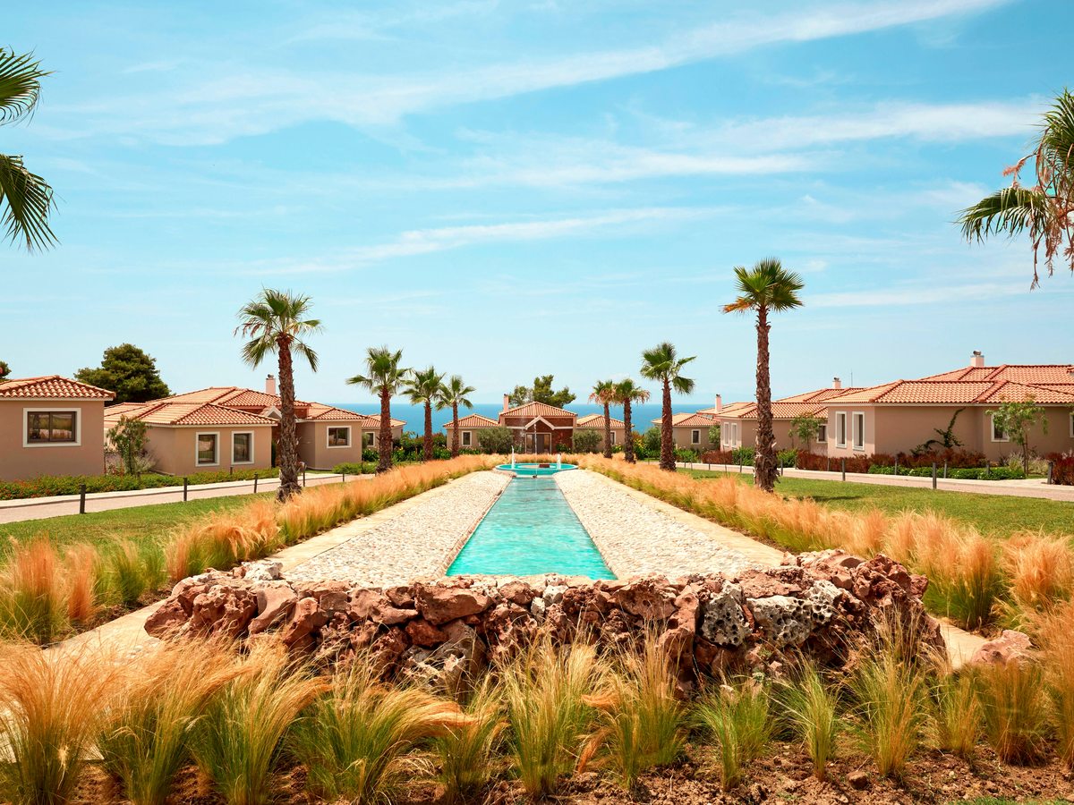 A long narrow ornamental water channel running between single-storey bungalows with terracotta roofs, edged by tall golden grasses and palm trees, with the sea on the horizon.