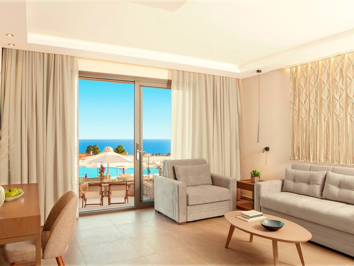 A suite living area with a grey sofa and armchair around a coffee table and a dining table by the window, with sliding doors onto a terrace above the pool and the sea.