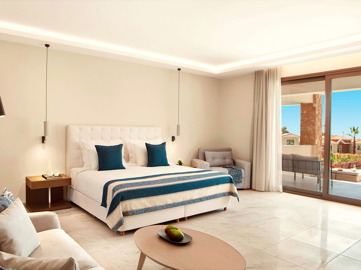 A large room with a double bed, a sofa and armchairs around a coffee table, pendant lights either side of the bed and doors onto a terrace.