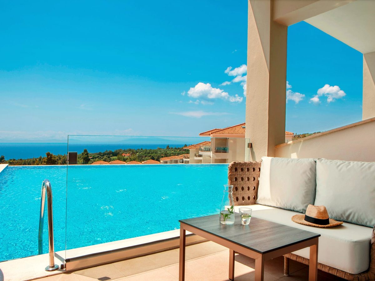 A shaded veranda with a cream sofa and a low table beside an infinity pool with a chrome ladder, looking over terracotta roofs to the sea.