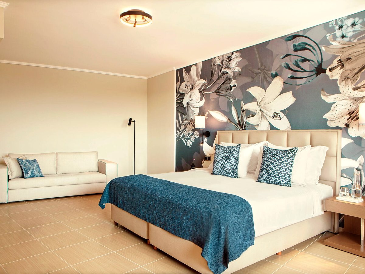 A large bedroom with a double bed under a grey-and-white floral mural, a teal throw across it and a cream sofa along the opposite wall.