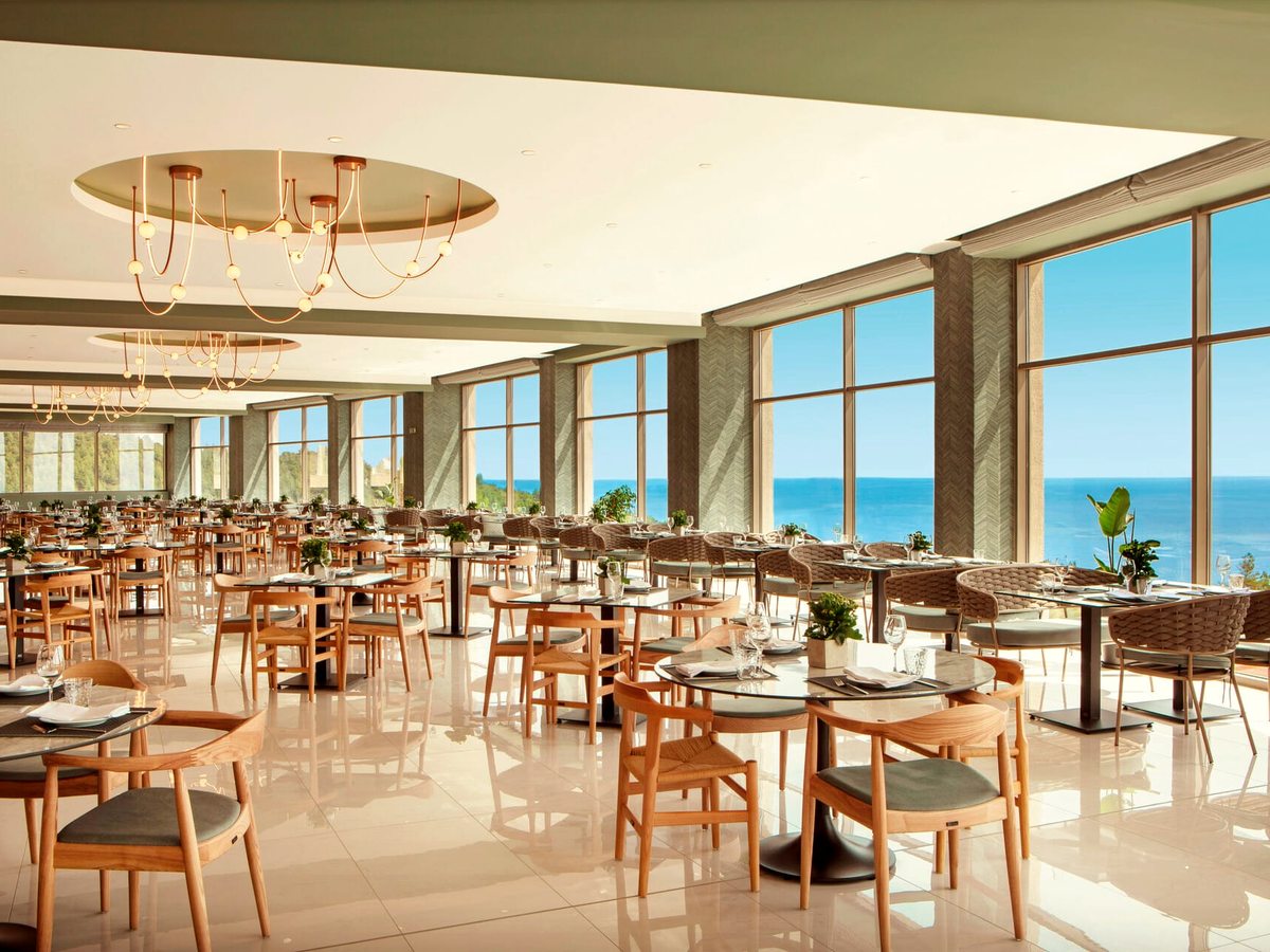 A large restaurant with pale wooden chairs at round and square tables, ring chandeliers overhead and full-height windows along one side looking out to the sea.