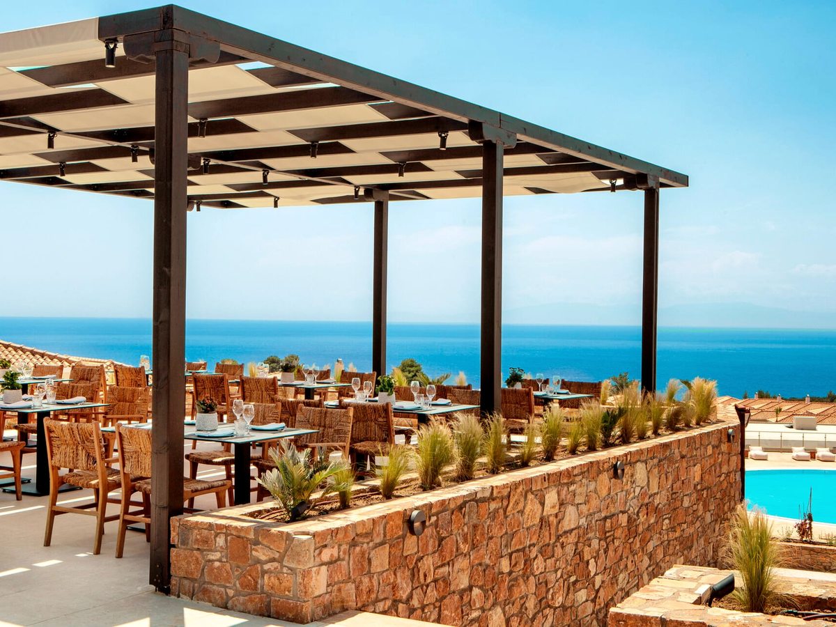 A dining terrace under a slatted pergola with woven wicker chairs at set tables behind a low stone wall, a pool to the right and the sea ahead.