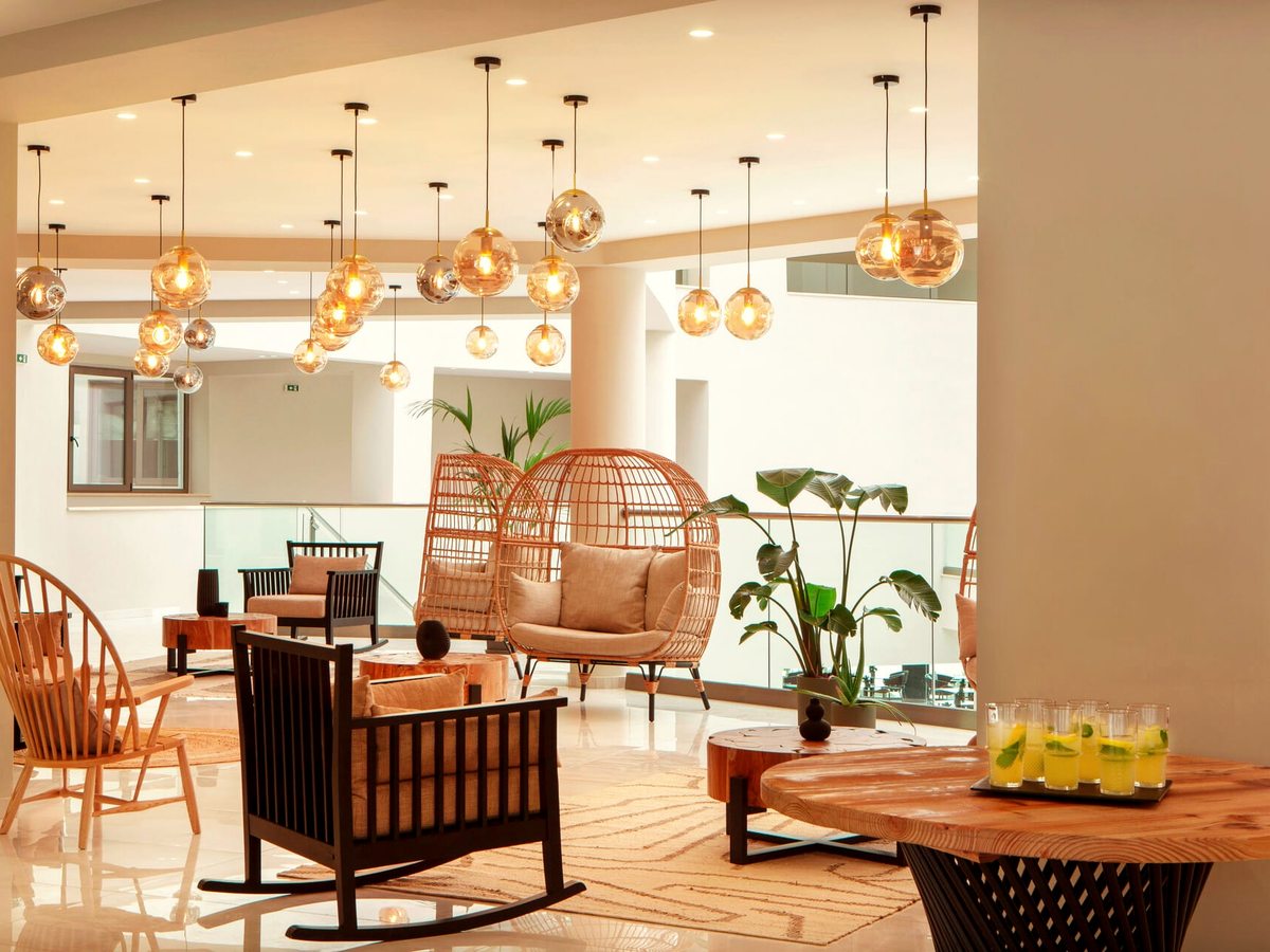 A hotel lounge with rattan hanging chairs, dark wooden rocking chairs, cream rugs and round wooden tables, under a cluster of amber glass pendant lights.