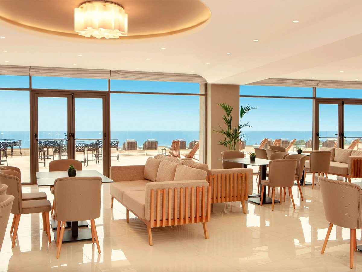 A lounge with cream armchairs and sofas at low tables, full-height glass doors onto a terrace of dark metal chairs and the sea beyond.