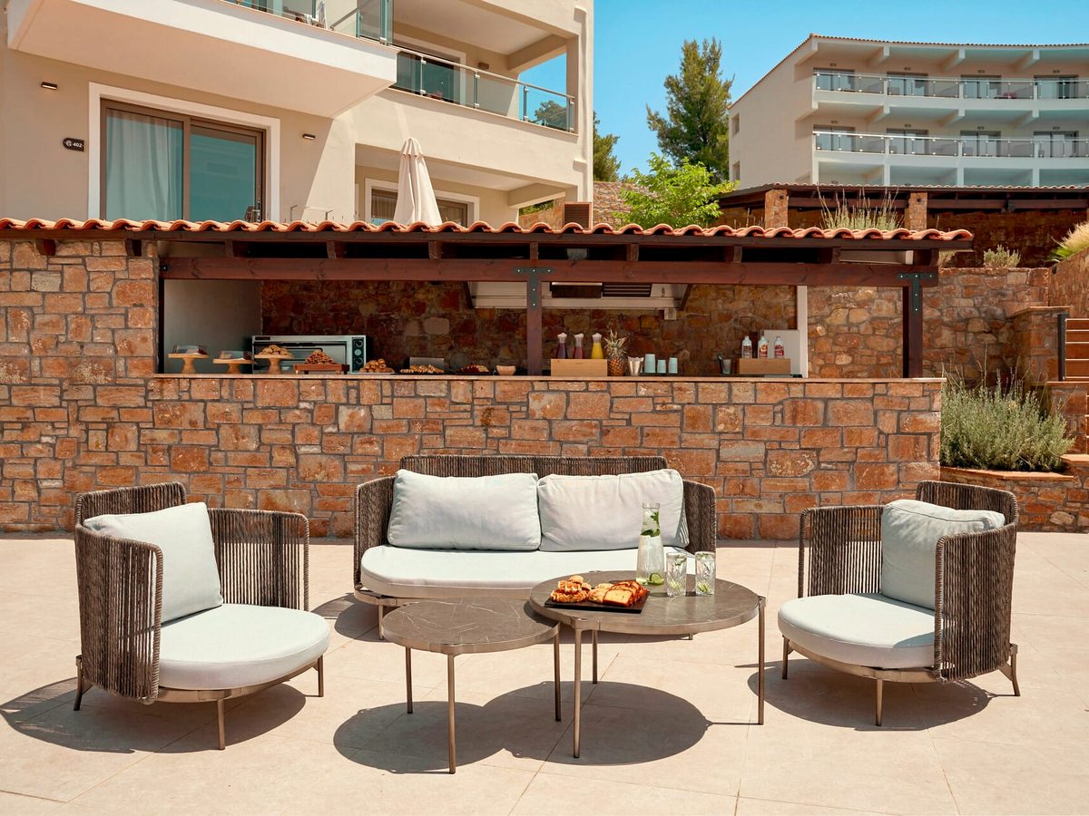 A stone-built outdoor counter with cakes, pastries and bottles laid out under a tiled canopy, with woven armchairs and a sofa around a coffee table in front of it.