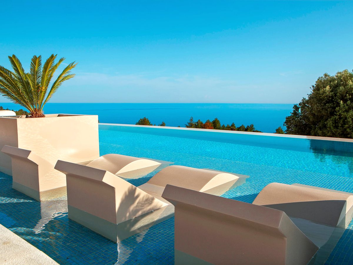 An infinity pool with cream in-water loungers along its near edge, a palm in a planter to one side and the sea beyond the far edge.