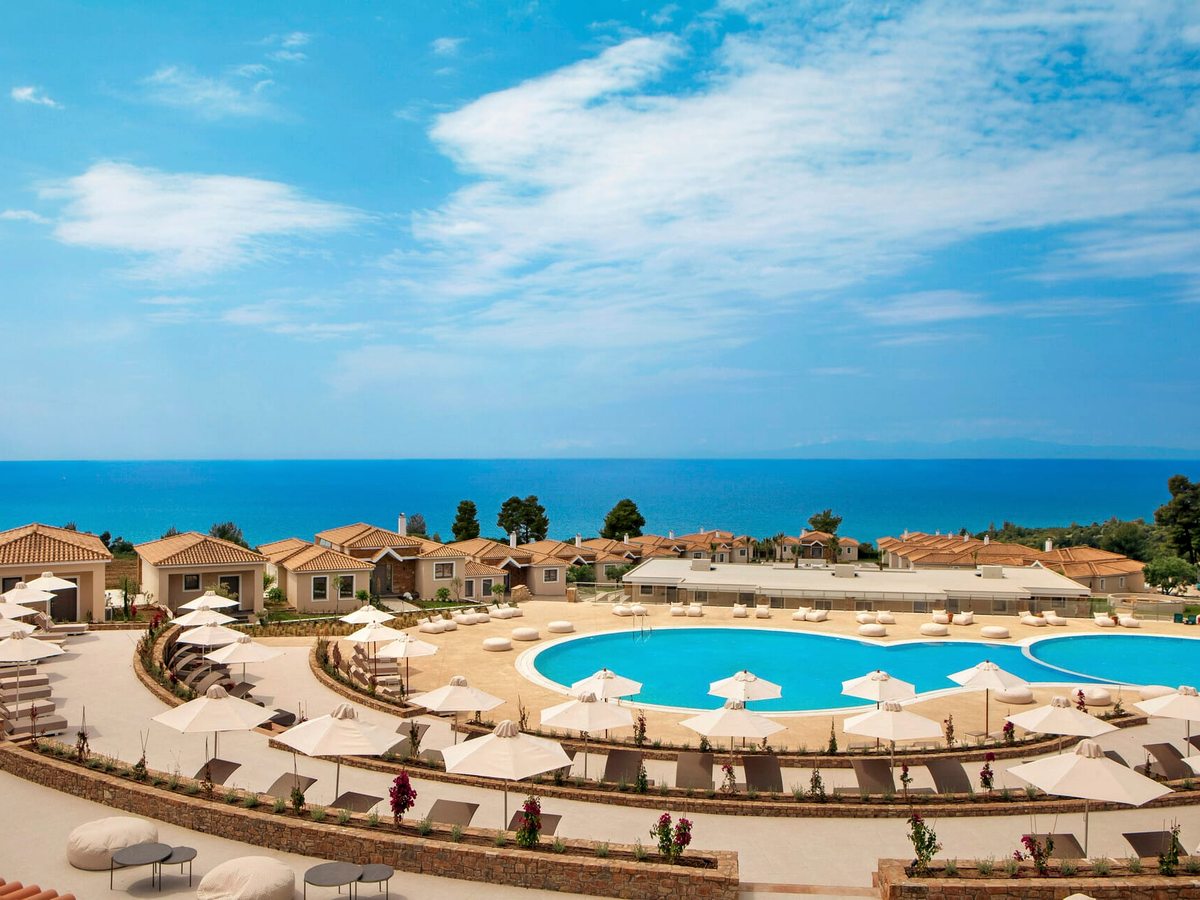 A large curved pool with rows of white parasols and sun loungers on stone terraces around it, bungalows and the sea behind.